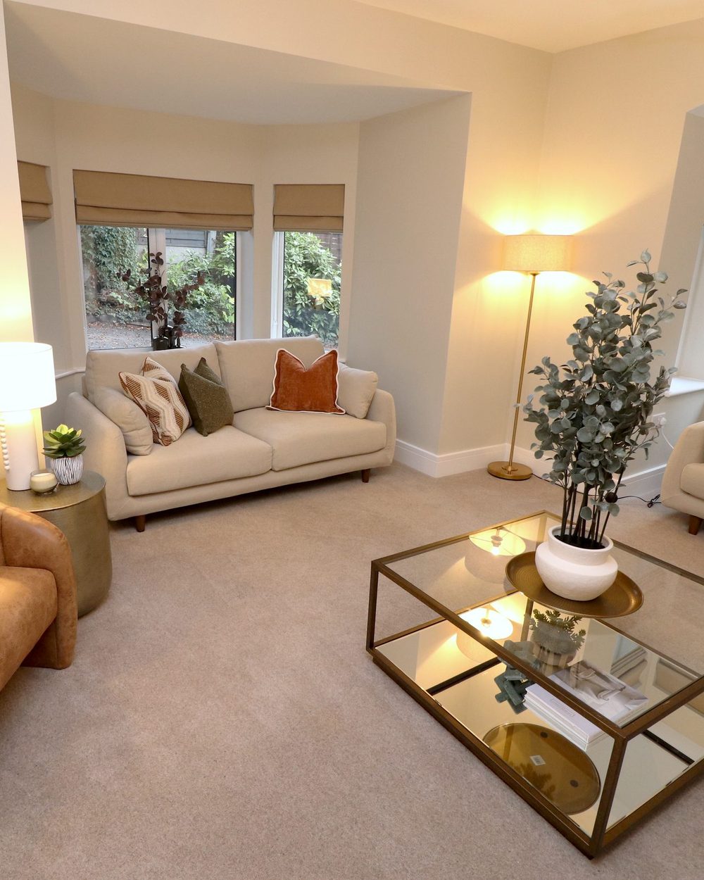 Roman blinds in a bay window above a cream sofa, floor lamp lit