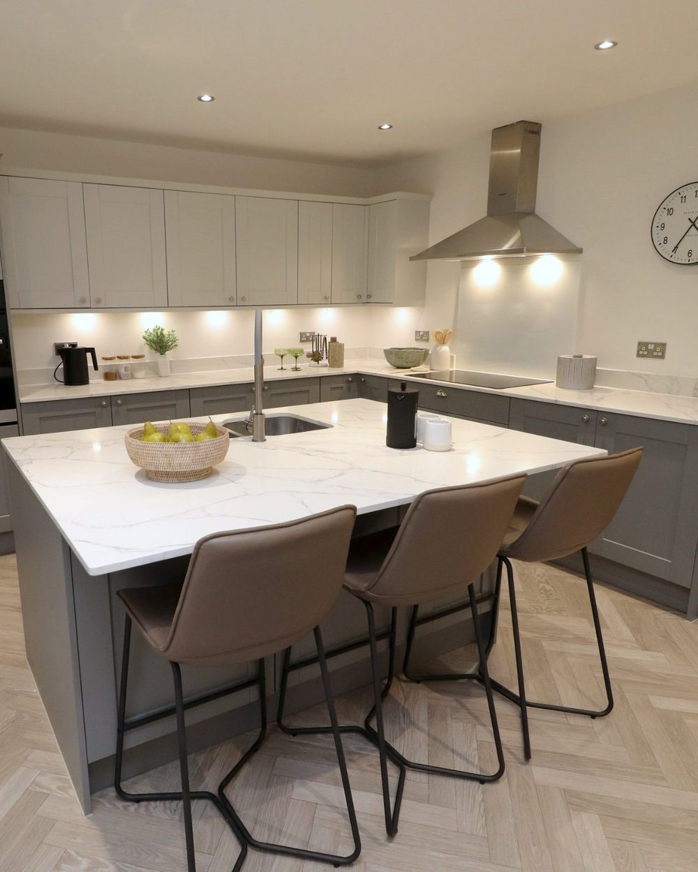 Staged kitchen: marble-topped island with three leather bar stools, a bowl of pears, grey shaker units and a herringbone floor