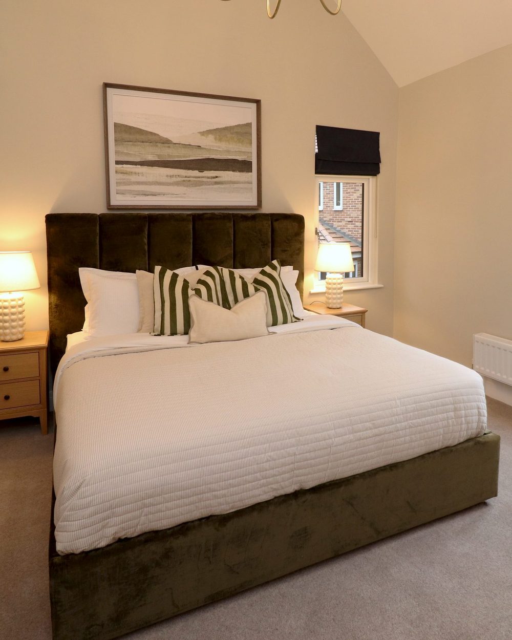 Staged bedroom: olive velvet bed, bedside lamp and a window with a blind