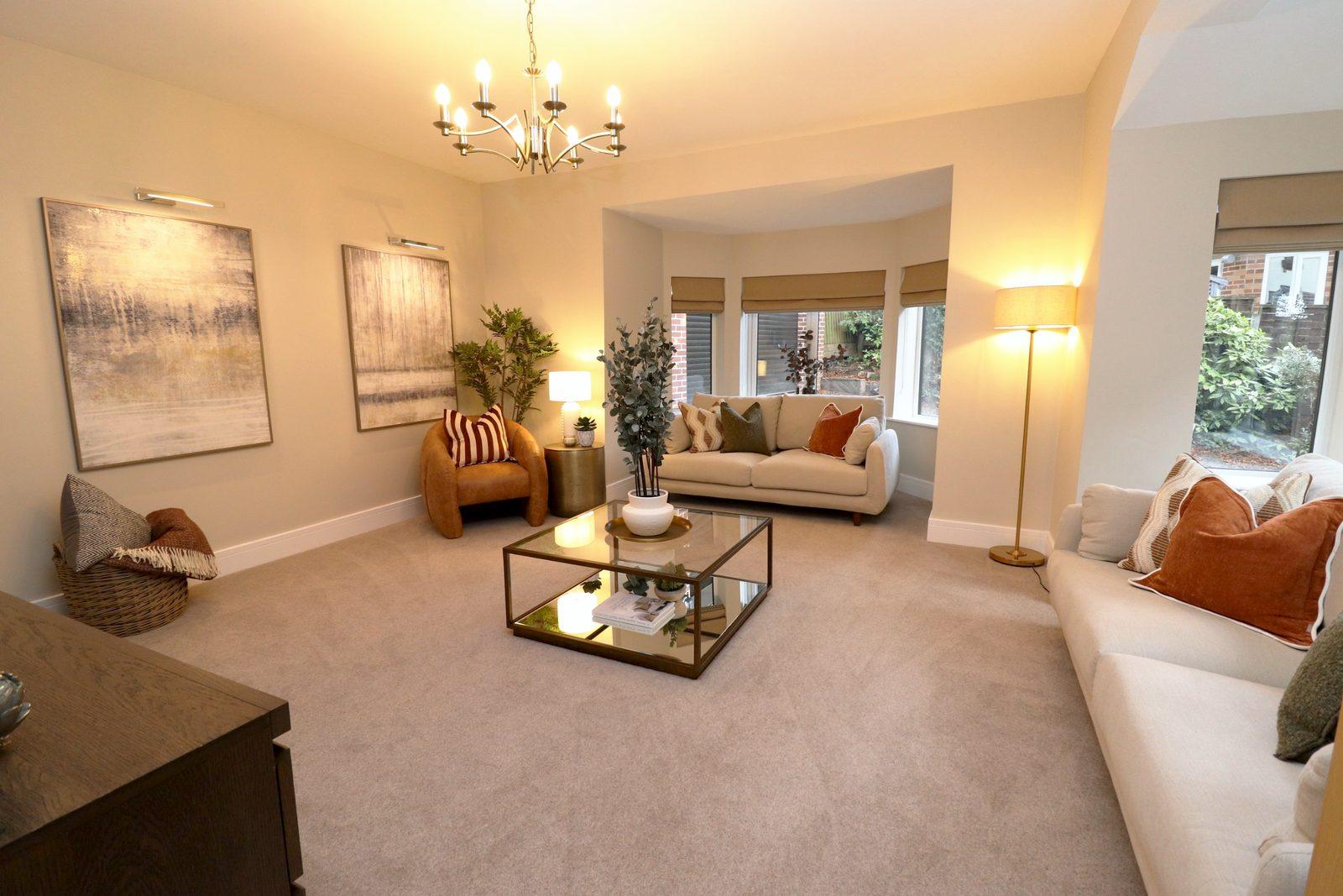 Living room after staging, from the door: two cream sofas and a tan leather armchair around a glass coffee table, two abstract canvases, a brass chandelier and the bay window beyond, lamps lit